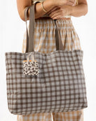 Eine Person, die eine hellbraune Karohose trägt, hält eine große braune Karotasche mit beigen Henkeln in der Hand. An der Tasche ist das „Aloha Tiny Tripper“ von Aloha Collection befestigt, ein kleines Accessoire mit Leopardenmuster. Von der Person sind nur der Oberkörper und der Arm zu sehen.