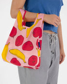 Eine Person mit blauem Top und grauer Hose hält die umweltfreundliche BAGGU Baby Tragetasche aus recyceltem Nylon mit bunten Apfel- und Bananenmotiven, kurzen Henkeln und farbenfrohem Design von BAGGU.