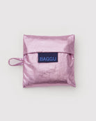 Die BAGGU Baby Tragetasche von BAGGU ist eine metallisch glänzende, pinkfarbene Tasche aus recyceltem Nylon mit einer Klappe, einer kleinen Seitenschlaufe und einem blauen "BAGGU"-Label auf der Vorderseite, das auf einem schlichten weißen Hintergrund abgebildet ist.