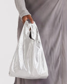 Eine Person in einem hellgrauen, langärmeligen Oberteil und einem seidengrauen Rock hält die BAGGU Baby Tragetasche, eine silbern glänzende Tasche aus recyceltem Nylon von BAGGU mit einem umweltfreundlichen, ausgeschnittenen Griffdesign.