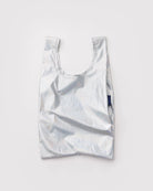 Eine zerknitterte, metallic-silberne BAGGU Baby Tragetasche aus recyceltem Nylon mit kurzen Henkeln wird vor einem weißen Hintergrund gezeigt, mit einem kleinen blauen Seitenanhänger, der das umweltbewusste BAGGU Design hervorhebt.