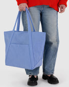 Eine Person mit rotem Pullover, blauen Jeans und schwarzen Schuhen hält die BAGGU Cloud Bag - eine große, umweltfreundliche Tasche von BAGGU aus recyceltem Nylon mit Doppelgriffen - vor einem unifarbenen Hintergrund.