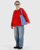 Eine Person mit kurzen grauen Haaren, Sonnenbrille, rotem Pullover, Kragenhemd, blauen Jeans und schwarzen Schuhen steht mit einer großen hellblauen BAGGU Cloud Bag von BAGGU vor einem schlichten hellen Hintergrund.