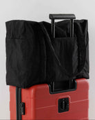 Eine große BAGGU Cloud Carry-On Tasche in Schwarz aus recyceltem Nylon sitzt auf einem roten Rollkoffer mit Griffen über dem Koffergriff. Schlichter weißer Hintergrund - perfekt als Handgepäck von BAGGU für unterwegs.