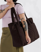 Eine Person in einem hellvioletten Oberteil und olivgrünem Rock trägt eine große BAGGU Cloud Carry-On Tasche aus recyceltem Nylon über der Schulter und zeigt tätowierte Arme. Der Hintergrund ist schlicht und hell.