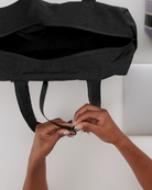 Man sieht die Hände einer Person, die die Riemen einer schwarzen BAGGU Everyday Cloud Bag von BAGGU zubindet, die teilweise geöffnet ist und auf einer weißen Fläche liegt.