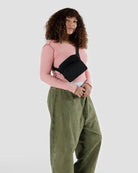 Eine Frau mit lockigem Haar trägt ein rosafarbenes langärmeliges Oberteil, eine weite olivgrüne Hose und die BAGGU Gürteltasche von BAGGU, die aus recyceltem Nylon besteht, während sie vor einem schlichten hellgrauen Hintergrund steht.