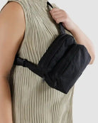 Eine Person in einem beigen Plissee-Oberteil trägt eine schwarze BAGGU-Gürteltasche aus recyceltem Nylon mit zwei Reißverschlussfächern, die sie sich quer über die Brust schnallt.