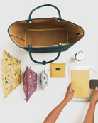 Eine Person legt einen gelben Beutel, eine Geldbörse und drei mit Blumenmustern bedruckte Reißverschlusstaschen neben eine geöffnete „BAGGU Large Nylon Reversible Tote“ und eine Wasserflasche auf eine weiße Fläche.