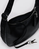 Die BAGGU Medium Recycled Leather Crescent Bag in Schwarz verfügt über einen breiten, geprägten "BAGGU"-Riemen, eine offene Reißverschlusstasche und eine abnehmbare Schlüsselleine mit zwei Schlüsseln, die vor einem hellen Hintergrund angebracht sind.
