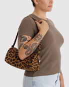Eine Person in einem braunen T-Shirt und hellen Jeans hält die BAGGU Nylon Loaf Bag von BAGGU mit Leopardenmuster und zeigt ihren tätowierten Arm mit einem Schlangen- und Blumenmuster vor einem schlichten hellen Hintergrund.