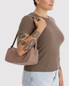 Eine Person in einem braunen T-Shirt und hellblauen Jeans trägt die BAGGU Nylon Loaf Bag von BAGGU auf der Schulter. Ihr tätowierter Arm zeigt ein Schlangen- und Sonnenmotiv vor einem schlichten weißen Hintergrund.