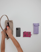 Jemand packt einen Gegenstand in eine beigefarbene BAGGU Nylon Loaf Bag von BAGGU. In der Nähe sind ein iPhone, ein rosafarbener Münzbeutel und ein lavendelfarbenes Brillenetui ordentlich auf einer weißen Fläche angeordnet.