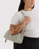 Eine Person mit Tätowierungen auf dem linken Arm, die ein weißes T-Shirt und eine schwarze Hose trägt, hält eine grün-weiße BAGGU Nylon Loaf Bag als Crossbody. Sie lächelt leicht und ist von den Schultern bis zur Taille abgebildet. Marke: BAGGU.