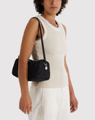 Eine Person in einem ärmellosen beigen Oberteil und einer weißen Hose steht vor einem schlichten Hintergrund und hält die „BAGGU Nylon Loaf Bag“ in der Hand – eine elegante schwarze Tasche aus recyceltem Material von BAGGU mit einem silbernen Anhänger.
