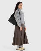 Eine Frau mit langen dunklen Haaren trägt eine graue Jacke, einen braunen Maxirock, gelbe Turnschuhe und graue Socken. Sie trägt eine BAGGU Nylon Shoulder Bag über der Schulter vor einem schlichten hellen Hintergrund.