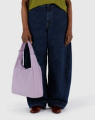 Eine Person steht vor einem hellen Hintergrund und zeigt nur ihren Unterkörper in einer blauen Jeans mit weitem Bein, einem grünen Oberteil und braunen Schuhen, während sie eine pastellviolette BAGGU-Nylon-Schultertasche von BAGGU hält.