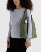 Eine Frau mit lockigem Haar, die teilweise außerhalb des Bildausschnitts zu sehen ist, trägt die BAGGU Nylon-Umhängetasche von BAGGU über der Schulter. Sie trägt ein hellgraues Langarmshirt und eine schwarze Hose und steht vor einem schlichten, hellen Hintergrund.