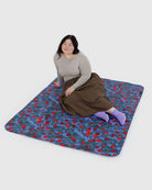 Eine Person mit langen dunklen Haaren, einem grauen Oberteil, einem braunen Rock und lavendelfarbenen Socken sitzt auf der BAGGU Puffy Picnic Blanket von BAGGU, die ein blaues, rotes und grünes botanisches Muster auf einem schlichten weißen Hintergrund zeigt.