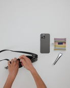 Jemand öffnet eine schwarze BAGGU Recycled Leather Pochette auf einer weißen Fläche neben einem Smartphone, einem gestreiften BAGGU-Beutel und einer silbernen Tube, möglicherweise Handcreme.
