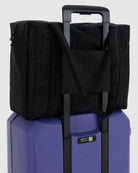 Eine BAGGU Small Cloud Carry-On Tasche aus schwarzem, recyceltem Nylon ist auf einem lilafarbenen Hartschalenkoffer mit verlängertem Griff befestigt, dessen Gurte über den Koffer drapiert sind - perfekt als BAGGU Untersitz-Reisetasche.