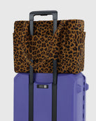 Die BAGGU Small Cloud Carry-On Tasche aus recyceltem Nylon liegt auf einem lilafarbenen Hartschalenkoffer, die Henkel über dem ausgezogenen Griff. Ideal als 16-Zoll-Laptoptasche oder praktisch unter dem Flugzeugsitz.