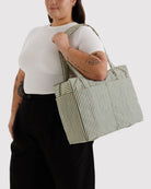 Eine tätowierte Person, die ein weißes kurzärmeliges Hemd und eine schwarze Hose trägt, hält die BAGGU Small Cloud Carry-On Tasche von BAGGU auf ihrer Schulter vor einem schlichten hellen Hintergrund.