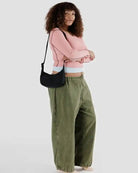 Eine Person mit lockigem Haar steht seitlich, trägt ein pinkfarbenes langärmeliges Oberteil, eine weite grüne Hose und trägt die BAGGU Small Nylon Crescent Bag von BAGGU vor einem schlichten hellen Hintergrund.
