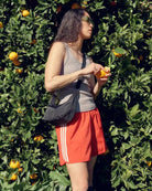Eine Frau mit Sonnenbrille, einem grauen Tanktop, roten Shorts und einer schwarzen BAGGU Small Nylon Crescent Bag steht vor einem Orangenbaum und schält und isst eine Orange, während das Sonnenlicht sie und die grünen Blätter beleuchtet.