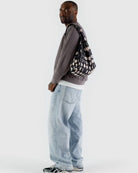 Ein Mann in grauem Strukturpullover, weißem T-Shirt, hellblauen Baggy-Jeans und grauen Turnschuhen steht seitlich mit einer BAGGU Standard Tragetasche von BAGGU aus recyceltem Ripstop-Nylon vor einem schlichten hellen Hintergrund.