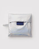 Die BAGGU Standard Tragetasche von BAGGU besteht aus recyceltem silbernem Ripstop-Nylon, ist faltbar, hat einen Flap-Verschluss und eine seitliche Schlaufe. Vorn befindet sich ein blaues Rechtecketikett mit "BAGGU" in schwarzen Buchstaben.