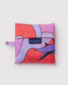 Die BAGGU Standard Tragetasche von BAGGU ist eine nachhaltige Einkaufstasche aus recyceltem Ripstop-Nylon mit buntem, abstraktem Muster in Rot, Lila und Lavendel. Sie kommt gefaltet in einer Hülle mit Seitenloop und blauem BAGGU Label.