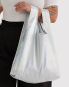 Eine Person mit Tätowierungen auf dem Arm hält eine große, glänzende BAGGU Standard Tragetasche aus recyceltem Ripstop-Nylon mit breiten Henkeln. Sie trägt ein weißes Hemd und eine schwarze Hose. Die Tasche ist von BAGGU.