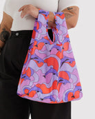 Eine Person mit schwarzen Hosen und weißem Shirt trägt die BAGGU Standard Tragetasche aus recyceltem Ripstop-Nylon mit abstraktem Muster und Delfinen von BAGGU; die Tattoos am Arm sind sichtbar.
