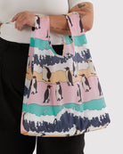 Eine Person in weißem Shirt und schwarzer Hose hält die BAGGU Standard Tragetasche von BAGGU aus recyceltem Ripstop-Nylon mit Pinguin- und Wellenmuster in Pink, Teal, Gelb und Navy. Tattoos sind am Arm sichtbar.