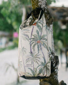 Ein wasserdichter Rucksack mit tropischem Palmenmotiv hängt an einem Baumstamm. Der Rucksack hat einen weißen Hintergrund und ein Etikett mit der Aufschrift „Meraki Supply Co.“ Der Baumstamm ist mit weißem Sand bedeckt und im Hintergrund sind üppige grüne Blätter zu sehen.
