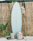 Ein hellblaues Surfbrett lehnt an einem Bambuszaun. Drei Main Design Dry Bag von Meraki Supply Co.-Taschen mit Palmenmotiven hängen am Zaun und liegen im Sand. Im Hintergrund sind üppige grüne Pflanzen und Bäume zu sehen.