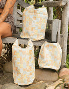 Drei Dry Bags von Meraki Supply Co. by Main Design mit geometrischen Mustern in Orange und Beige werden auf und neben einer rustikalen Bank im Freien mit Steinen, Grünpflanzen und einer teilweise sichtbaren Person in gemusterten Shorts ausgestellt.