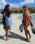 Ein lächelnder Mann und eine lächelnde Frau laufen barfuß mit Surfbrettern, Handtüchern und einem Dry Bag von Meraki Supply Co. von Main Design an einem Sandstrand entlang. Boote und Bäume prägen den Hintergrund unter einem klaren blauen Himmel.