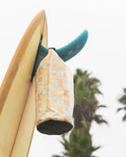 Die Dry Bag von Meraki Supply Co. by Main Design mit orangefarbenen und blaugrünen Mustern hängt an einer Surfbrettflosse mit Palmen und einem bewölkten Himmel im Hintergrund.