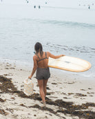 Eine Person in Badebekleidung und Shorts geht über einen mit Algen bedeckten Strand in Richtung Meer. Sie trägt ein Surfbrett und einen Dry Bag von Meraki Supply Co. von Main Design. Auf dem Wasser sind mehrere Surfer zu sehen.