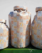 Drei große Dry Bag von Meraki Supply Co. by Main Design mit pastellfarbenen geometrischen und illustrativen Motiven – Wellen, Blumen, „SAGRATE“ – stehen aufrecht auf einer grünen Fläche. Die Taschen sind wasserdicht und stilvoll gemustert.