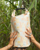 Eine Person mit mehreren Ringen hält im Freien zwischen hohen, grünen Pflanzen einen gemusterten Dry Bag von Meraki Supply Co. by Main Design über ihrem Kopf und hebt so seine stilvollen und wasserdichten Eigenschaften hervor.