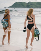 Zwei Frauen in Badeanzügen laufen barfuß über einen Sandstrand und tragen die Dry Bag von Meraki Supply Co. by Main Design mit tropischen Motiven. Im Hintergrund erscheinen Meereswellen und grüne Hügel unter einem teilweise bewölkten Himmel.