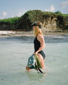 Eine Frau in schwarzem Badeanzug und Schnorchelmaske steht im flachen Meerwasser und hält Flossen und einen Dry Bag von Meraki Supply Co. by Main Design. Im Hintergrund sind felsige grüne Klippen und ein Sandstrand unter einem teilweise bewölkten Himmel zu sehen.