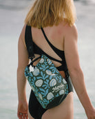 Eine Person in schwarzem Badeanzug steht am Wasser und trägt die wasserdichte Dry Bag von Meraki Supply Co. by Main Design mit blauem und grünem Blumenmuster auf dem Rücken.