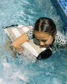 Ein junges Mädchen in einem gemusterten Badeanzug schwimmt in einem Freibad und schmiegt sich an ein gestreiftes aufblasbares Spielzeug, während das Wasser am gefliesten Rand plätschert - ihr Dry Bag von Meraki Supply Co. by Main Design liegt in der Nähe.