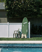 Ein grünes Surfbrett lehnt an einem weißen Zaun neben einem mit einem Handtuch drapierten Plastikstuhl. Vier wasserfeste Metallkanister und ein Main Design Dry Bag von Meraki Supply Co. stehen am blauen Pool - ideal für Ihr nächstes Outdoor-Abenteuer.