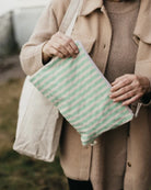 Eine Person in einem beigen Mantel hält eine große Tasche und benutzt die grün-weiß gestreifte Pocket Reissverschlusstasche von OMOM by Main Design als Organizer im Freien.