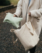 Eine Person in einem hellen, wuscheligen Mantel steht auf einem Kiesweg, in der Hand eine cremefarbene Bio-Baumwolle-Tasche und die Pocket Reissverschlusstasche von OMOM by Main Design in grün-weiss gestreift.